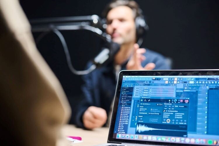 A man talking behind a podcast microphone with headphones on and a laptop with audio tracks in front of him. | © Plug The Jack