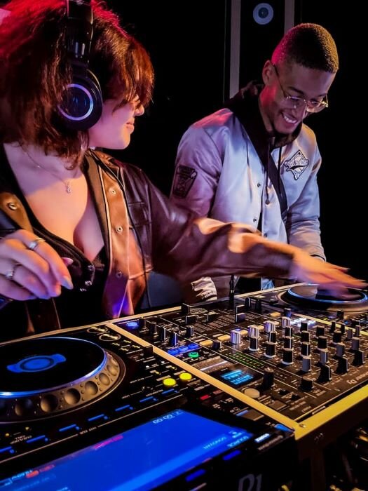 Couple à la table de mixage dans studio DJ, femme avec casque, homme regardant. | © Plug The Jack